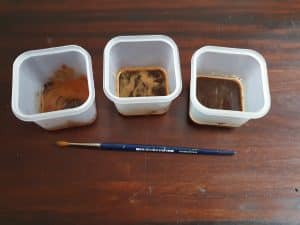 3 Plastic bowls with different strengths of coffee in each. A paintbrush in front of them on a wooden table.